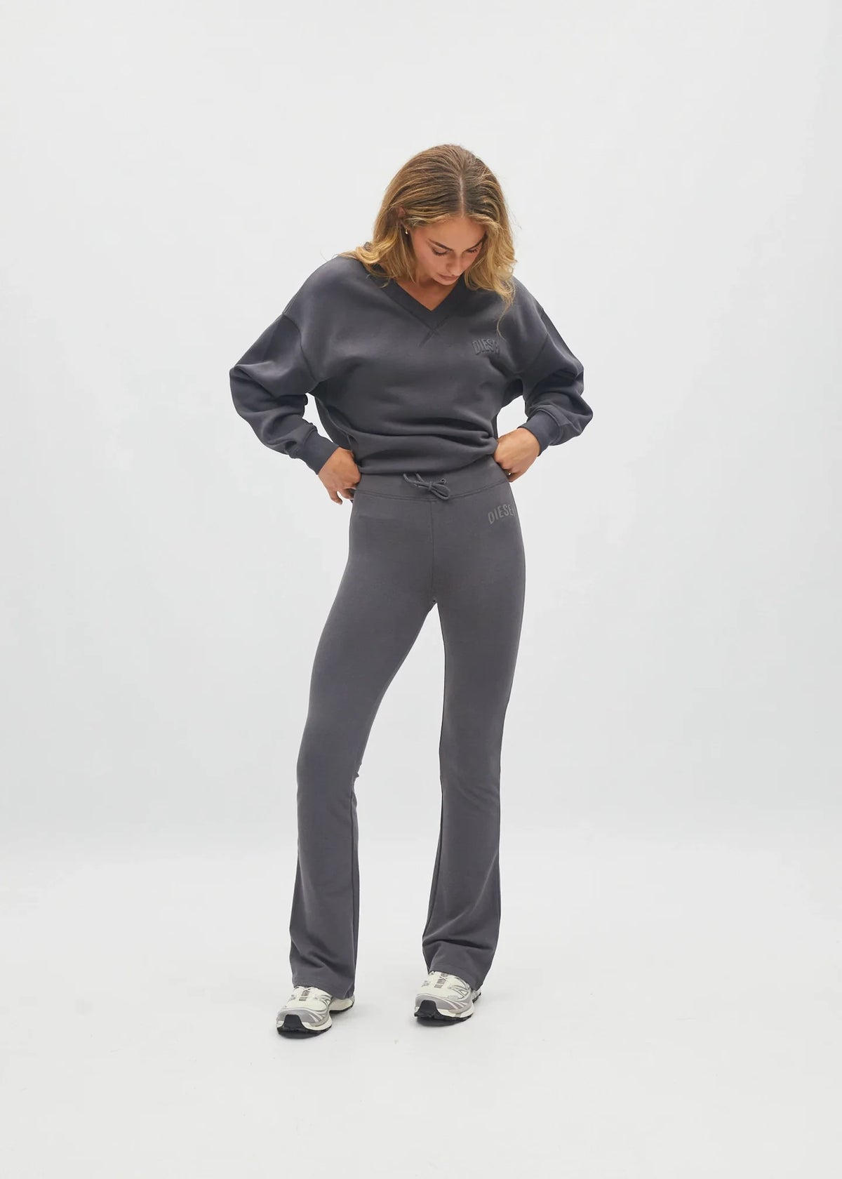 A woman modeling a dark gray lounge set consisting of a long-sleeve sweatshirt and matching flared sweatpants, standing against a light gray studio background.