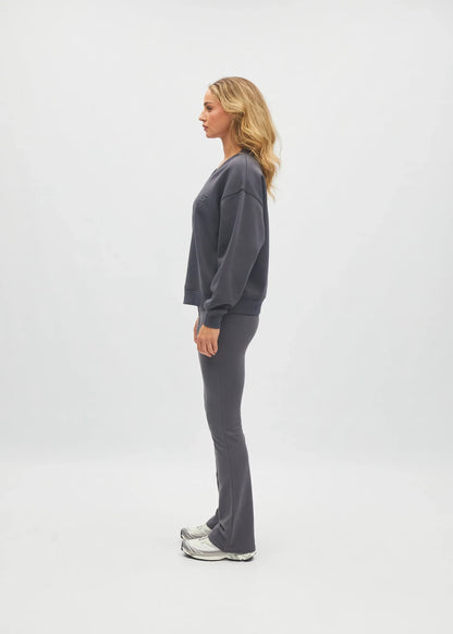 A woman stands in profile wearing a matching dark gray sweatshirt and jogger pants set, paired with white sneakers, against a plain light gray studio background.