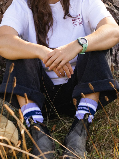 Person sitting outdoors wearing a white t-shirt, dark pants, and purple socks with 'Blubird' branding.