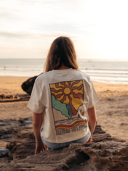 A woman sits on a rock at a beach, viewed from behind, wearing a cream-colored graphic t-shirt with a colorful retro-style sun and landscape print on the back.