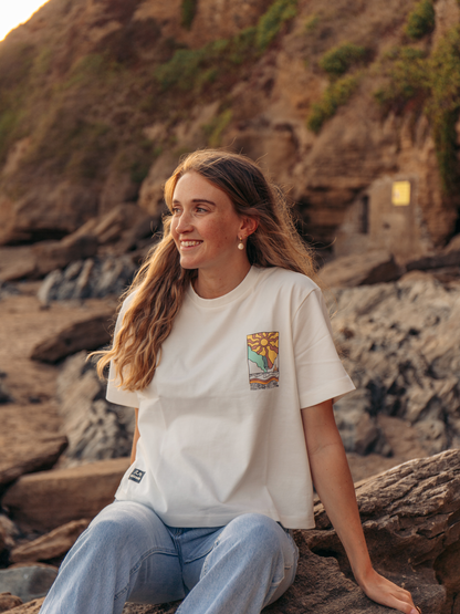 A woman sitting outdoors on a rocky cliffside wearing a white graphic t-shirt and light blue jeans, with warm golden-hour lighting.