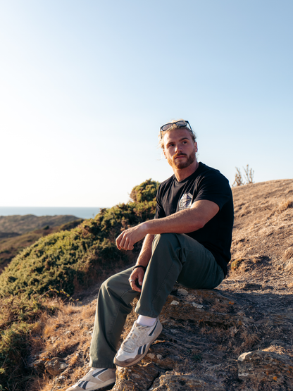 A man sitting outdoors on a rocky hillside under a clear blue sky, wearing a black graphic T-shirt, dark green pants, white sneakers, and sunglasses on his head.