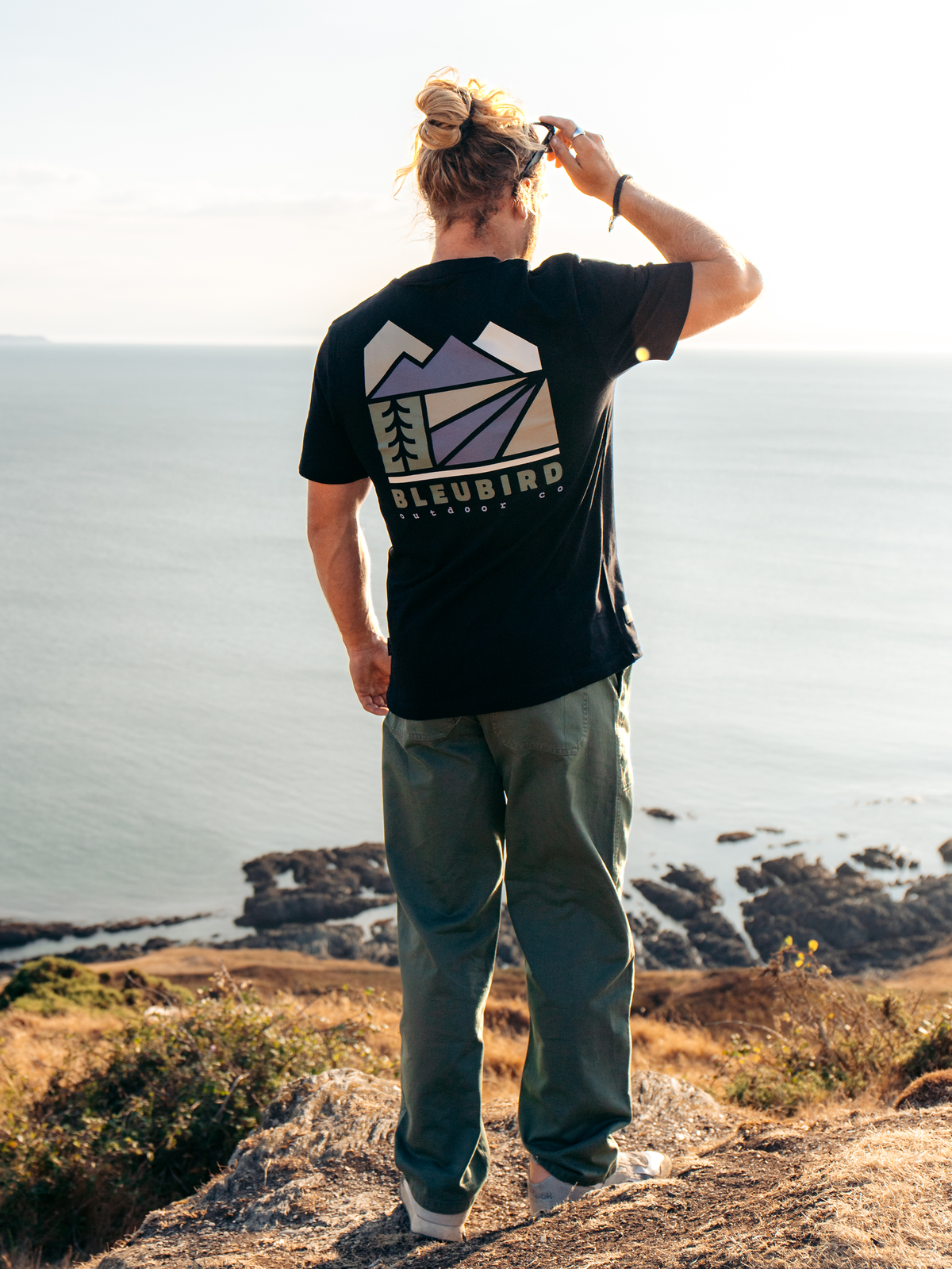 A man stands on a rocky cliff overlooking the ocean, wearing a black t-shirt with a large mountain graphic and the brand name 'BLEUBIRD' on the back, paired with olive green pants.