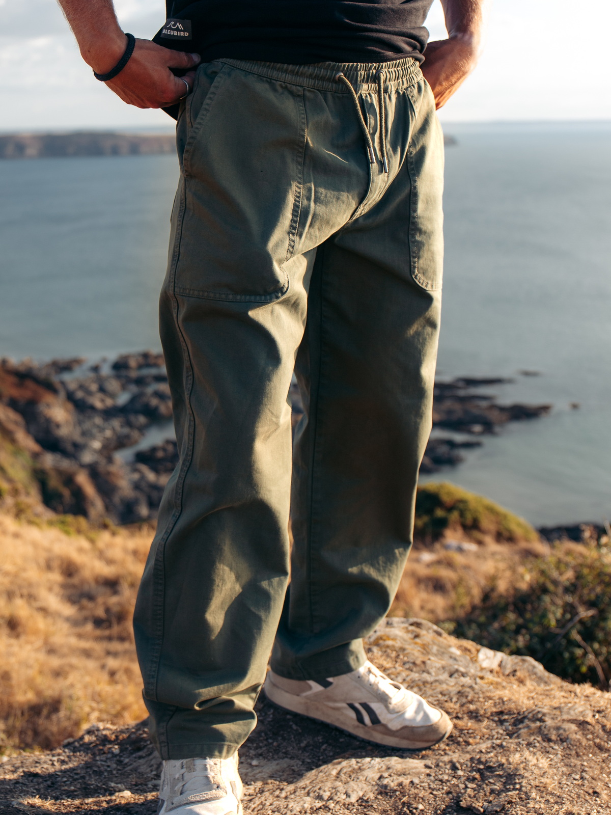 A person standing outdoors on a rocky cliff overlooking water, wearing loose olive green drawstring pants, a black top, and white sneakers.