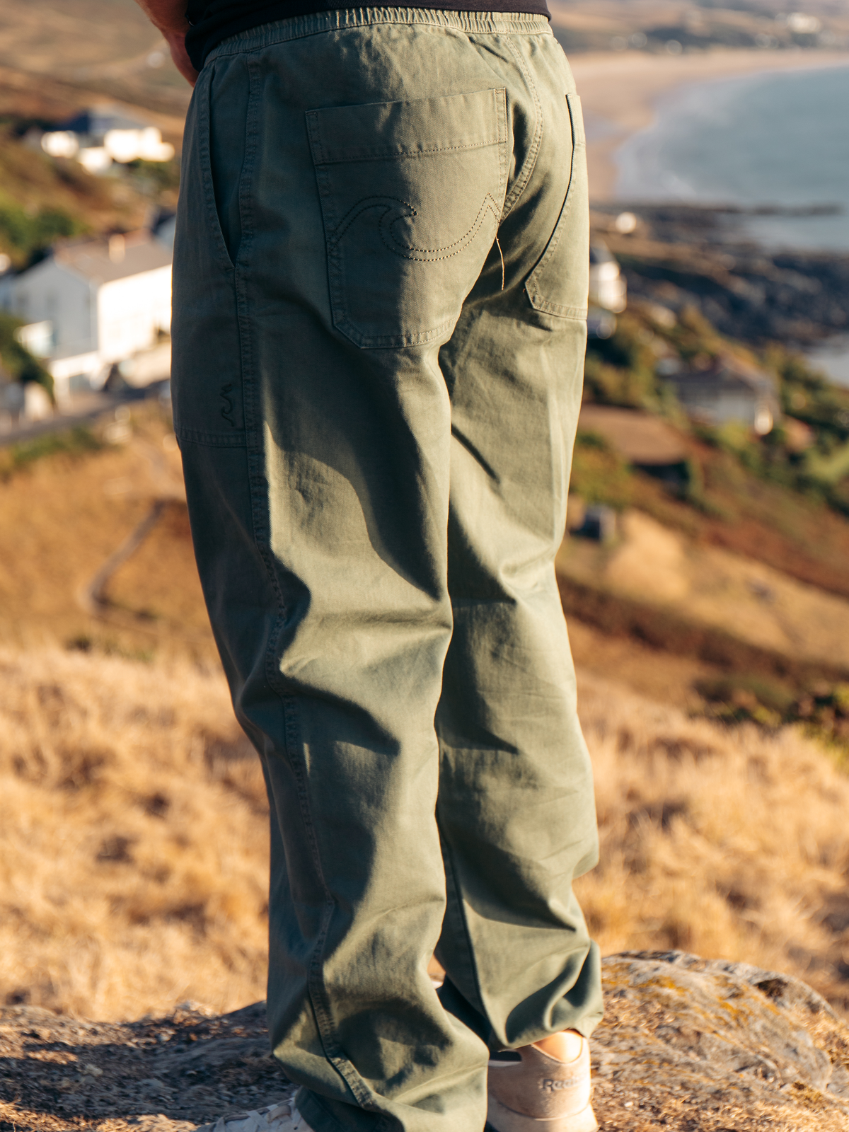 A person viewed from the back wearing olive green canvas pants with a large back pocket, standing outdoors on a rocky hillside overlooking a coastal landscape.