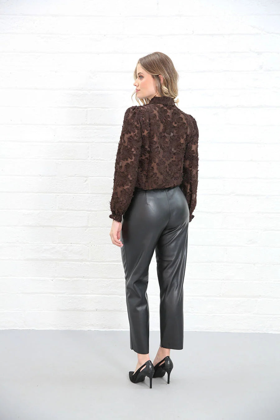 A woman is shown from the back wearing a brown patterned long-sleeve blouse and black faux-leather cropped pants, standing against a white brick wall.