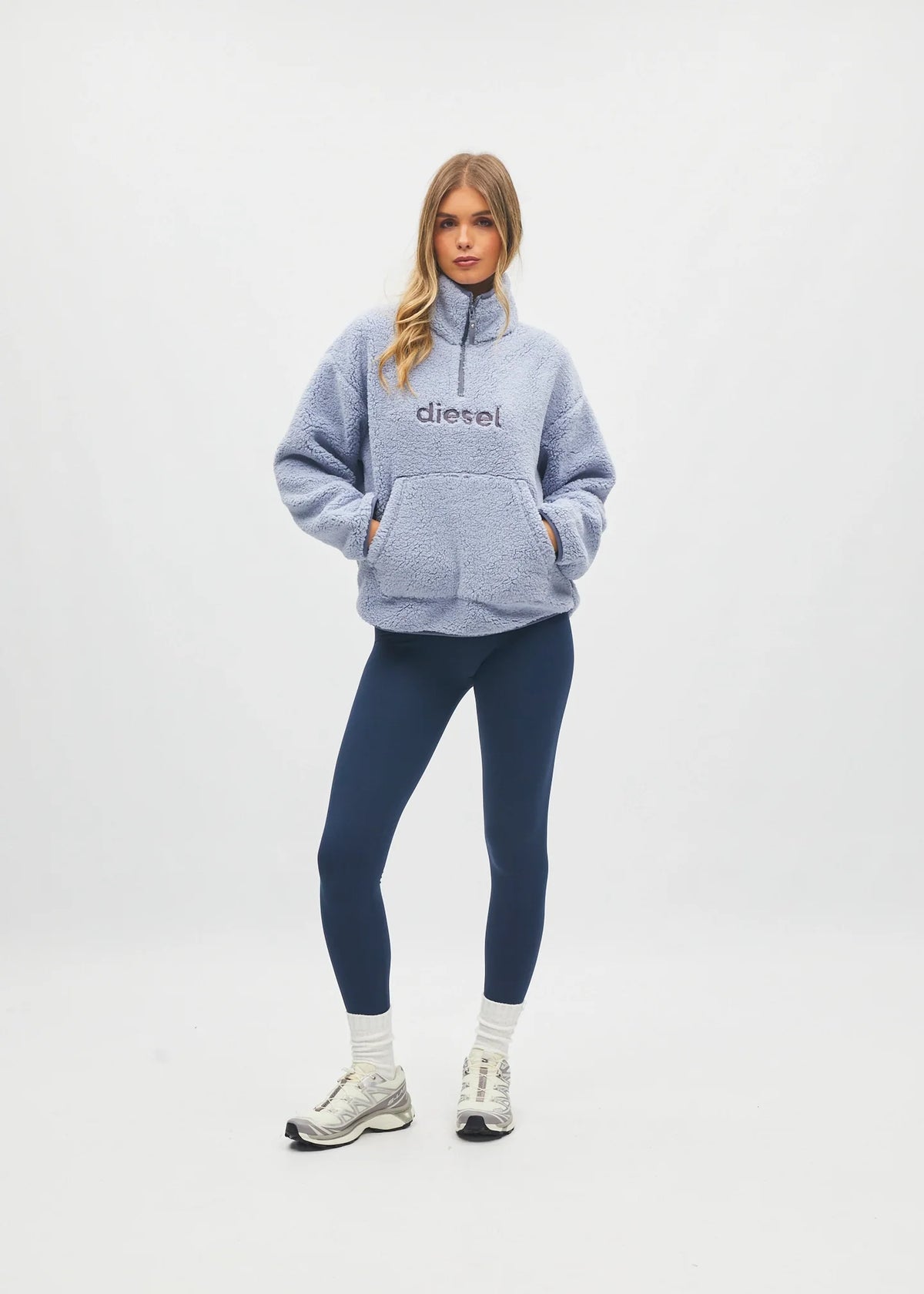 A woman modeling a light blue heathered half-zip fleece pullover sweatshirt with 'diesel' printed on the chest, paired with navy blue leggings, white socks, and white sneakers, against a plain white studio background.