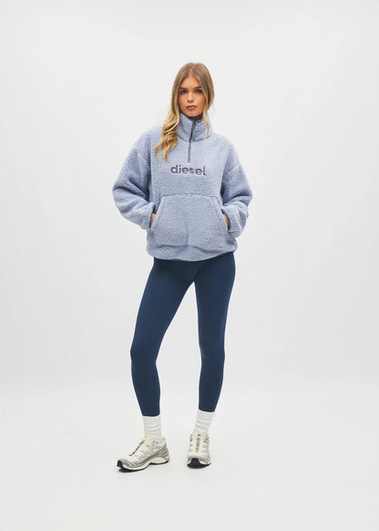 A woman modeling a light blue heathered half-zip fleece pullover sweatshirt with 'diesel' printed on the chest, paired with navy blue leggings, white socks, and white sneakers, against a plain white studio background.