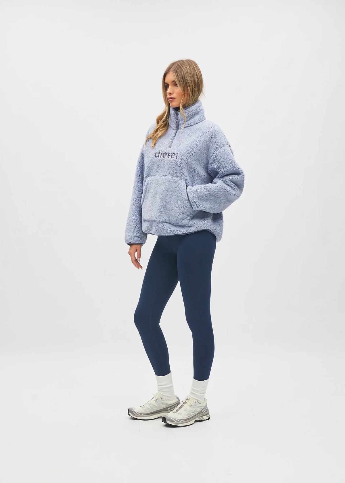 A woman modeling a light blue fuzzy quarter-zip pullover sweatshirt with 'c'chic!' embroidered on the chest, paired with navy blue leggings, white socks, and white sneakers, against a plain white studio background.