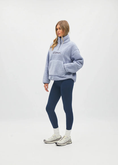 A woman modeling a light blue fuzzy quarter-zip pullover sweatshirt with 'c'chic!' embroidered on the chest, paired with navy blue leggings, white socks, and white sneakers, against a plain white studio background.