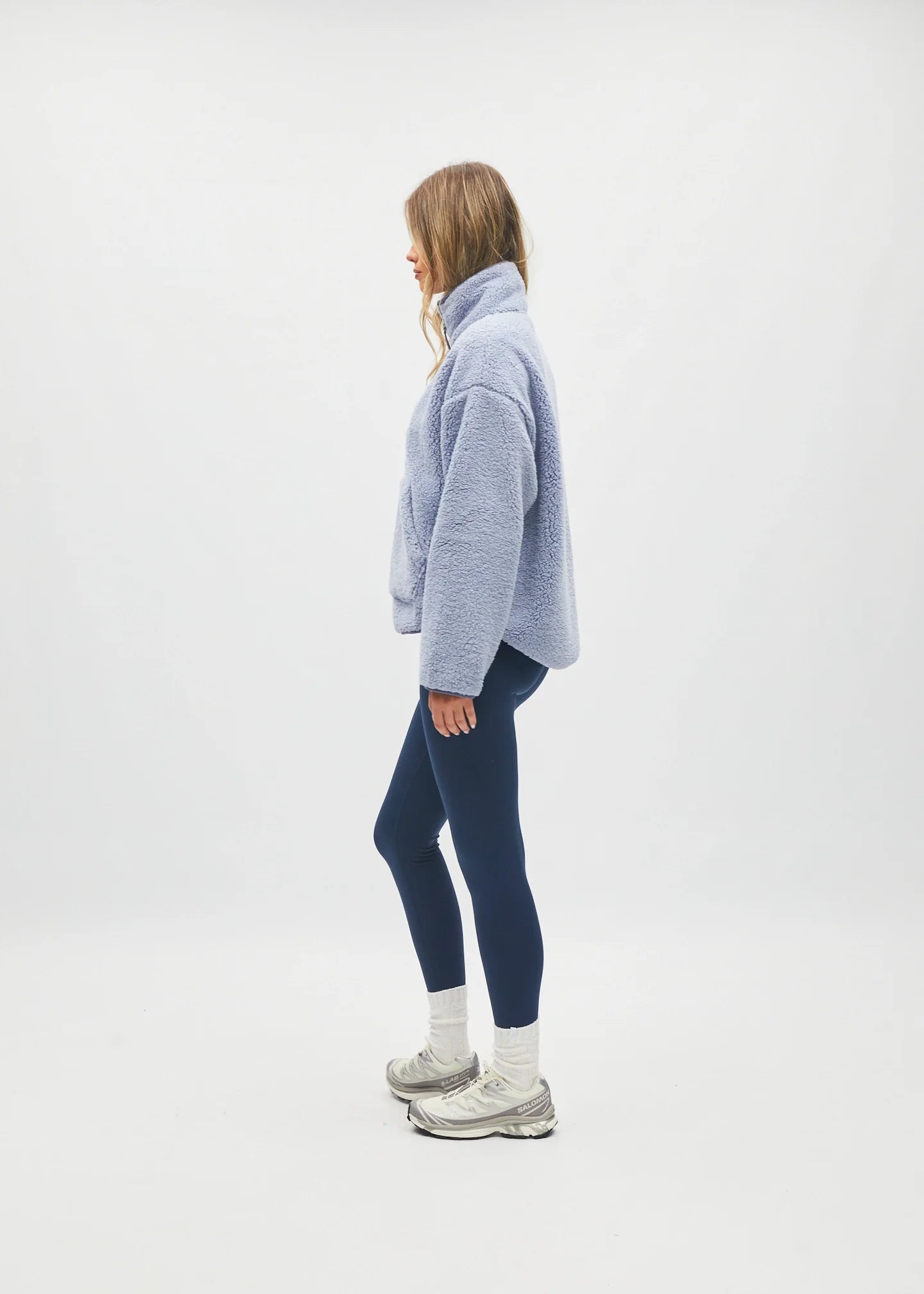 A woman stands in profile wearing a light periwinkle blue fuzzy cropped sweater, dark navy leggings, white socks, and gray sneakers against a seamless white studio background.