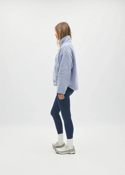 A woman stands in profile wearing a light periwinkle blue fuzzy cropped sweater, dark navy leggings, white socks, and gray sneakers against a seamless white studio background.