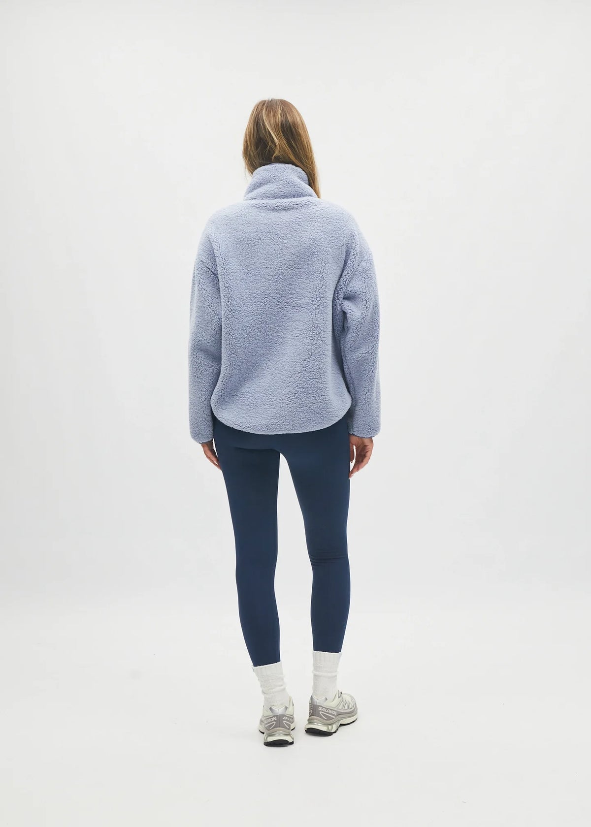 A woman is shown from behind wearing a light blue fuzzy fleece pullover, dark navy leggings, white socks, and gray sneakers, standing against a plain white studio background.