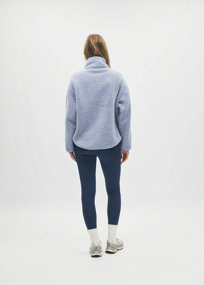 A woman is shown from behind wearing a light blue fuzzy fleece pullover, dark navy leggings, white socks, and gray sneakers, standing against a plain white studio background.