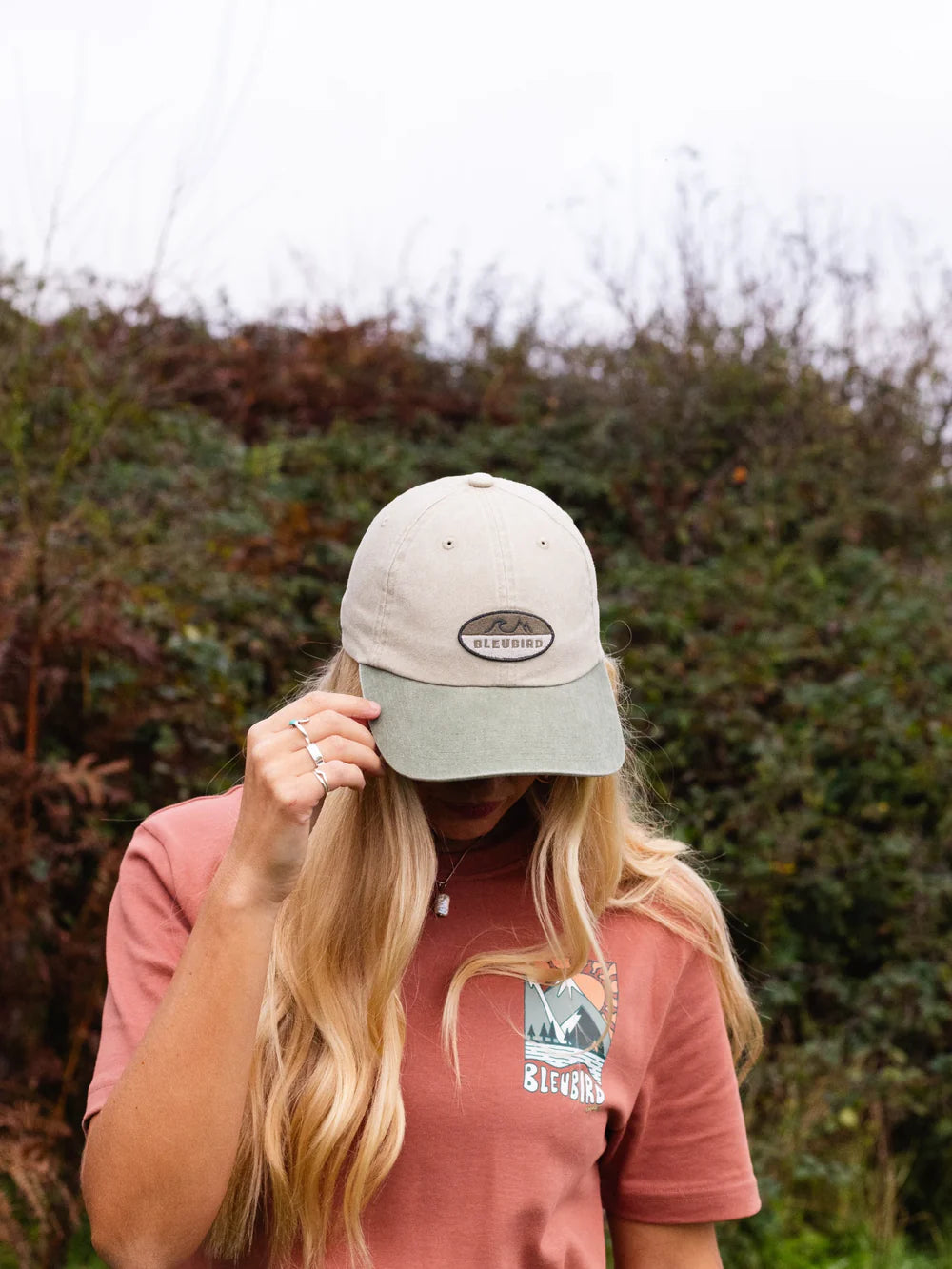 Person wearing a cap and t-shirt with a logo, standing in front of greenery