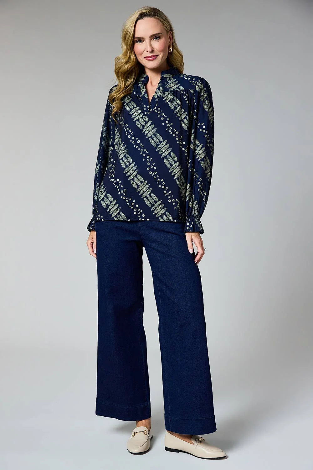 A woman modeling a navy blue long-sleeve button-up blouse with a beige geometric pattern, paired with navy blue wide-leg trousers and beige loafers, against a light gray studio background.