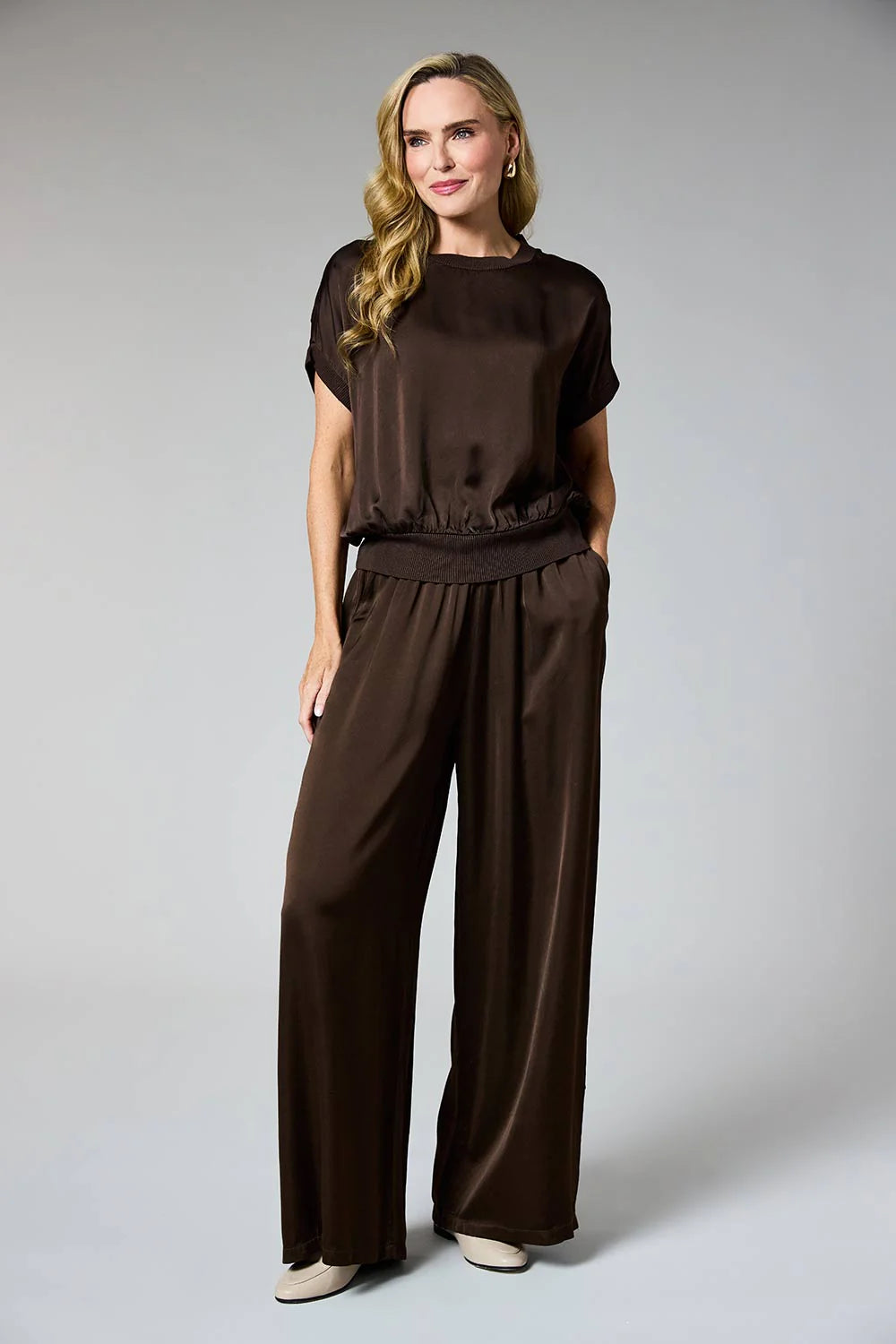 A woman modeling a dark brown satin jumpsuit with short sleeves, a gathered waist, and wide legs, photographed against a neutral gray studio background.