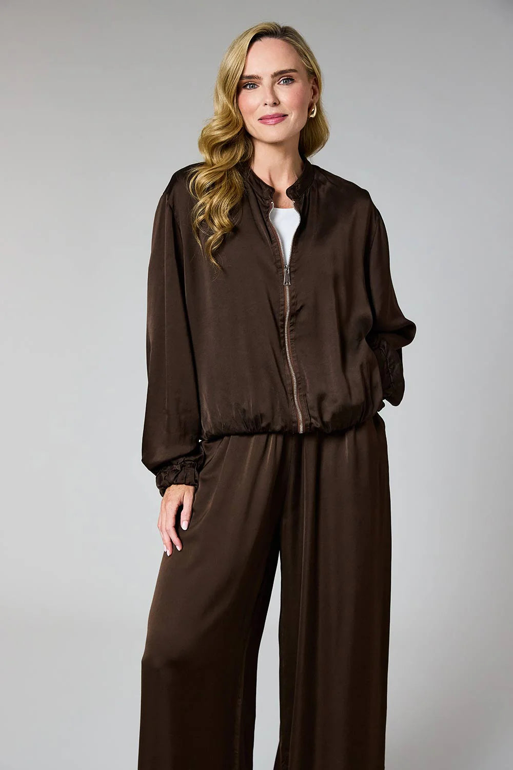 A woman modeling a dark brown satin-like jumpsuit with a zip-up jacket-style top and wide-leg pants against a plain light gray studio background.