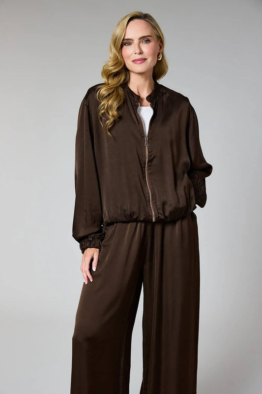 A woman modeling a dark brown satin-like jumpsuit with a zip-up jacket-style top and wide-leg pants against a plain light gray studio background.