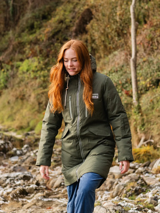 Person wearing a green jacket walking through a natural landscape with trees and rocks.