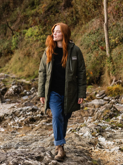 Woman with red hair wearing a green coat and blue jeans standing on a rocky path in a forest.