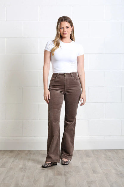 A woman modeling high-waisted brown corduroy flare pants with a white t-shirt, standing against a white brick wall and light wood floor.