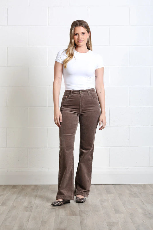 A woman modeling high-waisted brown corduroy flare pants with a white t-shirt, standing against a white brick wall and light wood floor.
