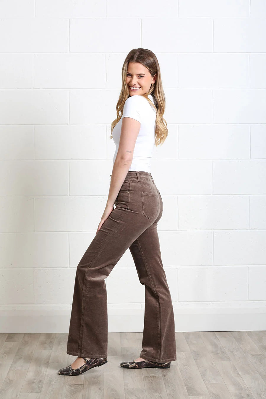 A woman modeling high-waisted brown corduroy flare pants, paired with a white t-shirt and dark flat shoes, standing against a white brick wall.