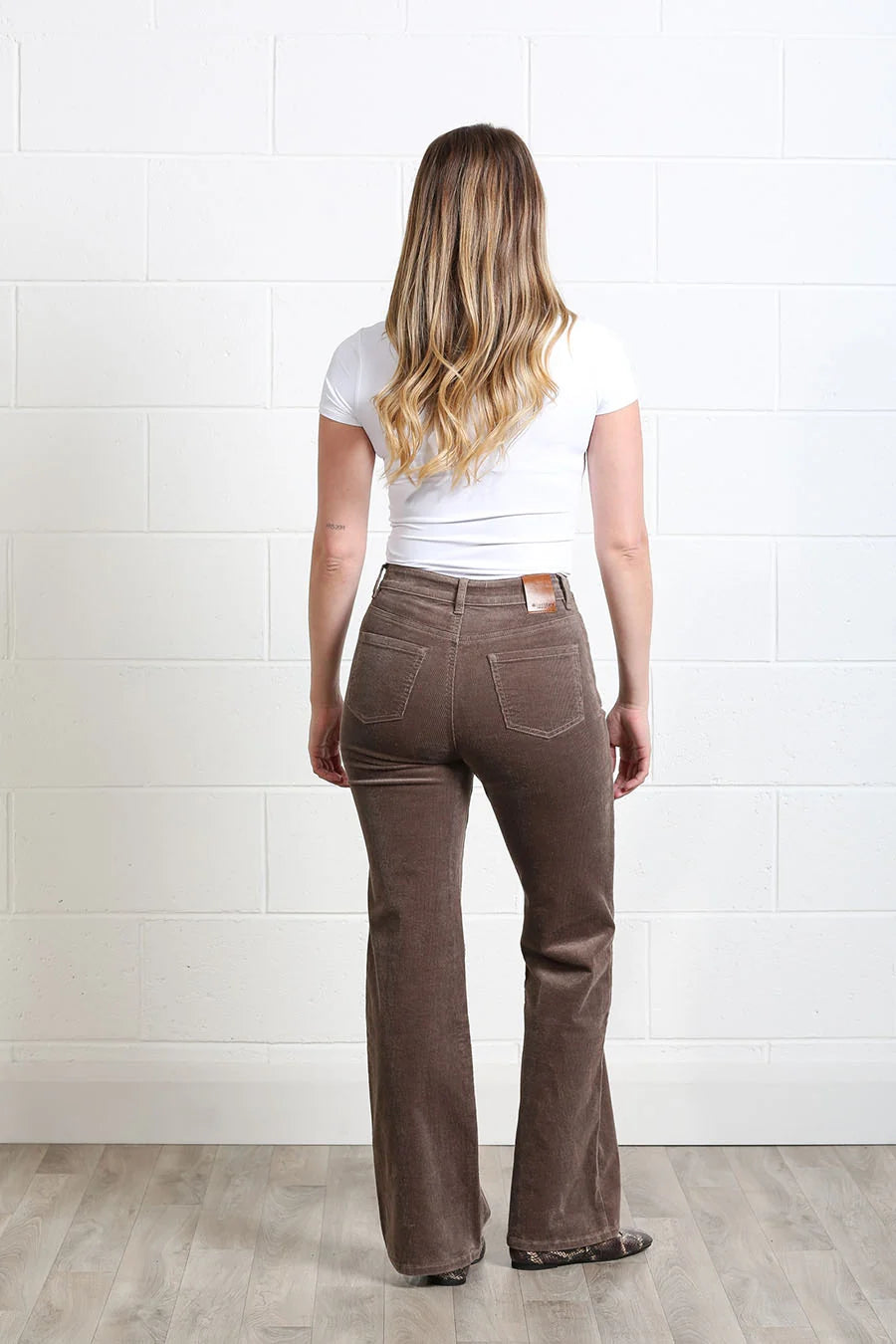 A woman viewed from behind wearing brown flared jeans, a white t-shirt, and black shoes, standing against a white brick wall and light wood floor.