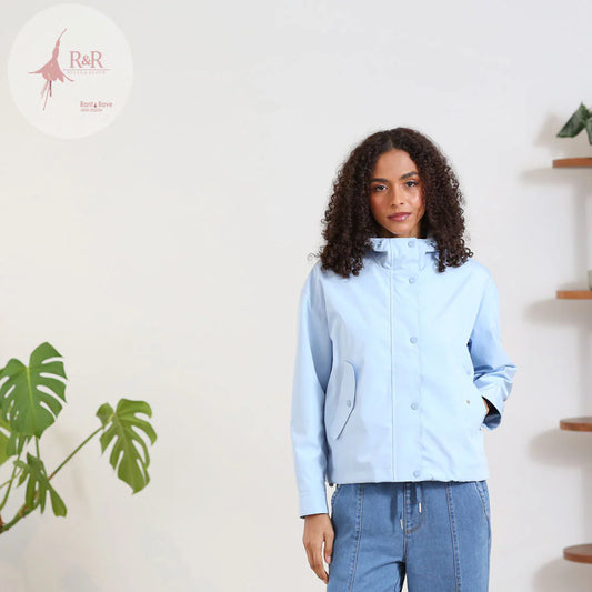 Woman wearing a light blue jacket standing in a room with a plant and shelves.