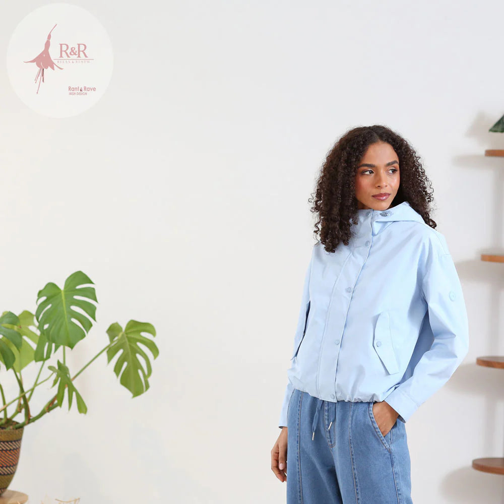 Woman wearing a light blue shirt and jeans standing in a room with a plant and shelves.