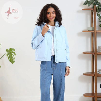 Woman wearing a light blue denim jacket and jeans standing in a room with a plant and wooden shelf.