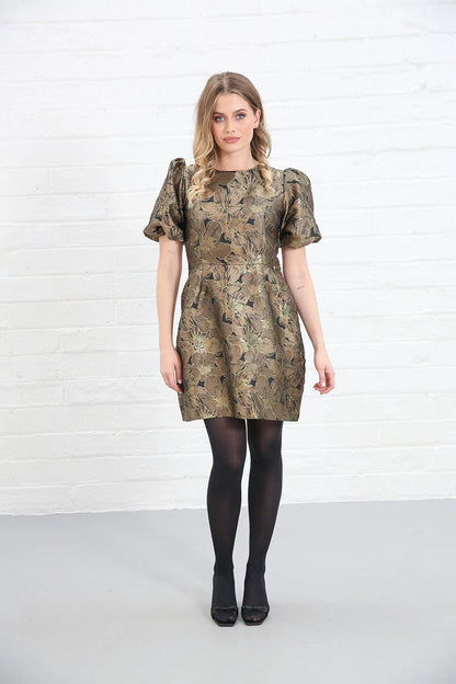 A woman modeling a short puff-sleeve dress with an abstract gold, brown, and black pattern, paired with black tights and black shoes, standing against a white brick wall.