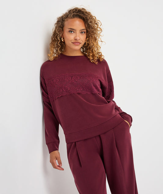 A woman with curly blonde hair models a matching burgundy lounge set consisting of a long-sleeve top with a lace chest panel and matching wide-leg pants, set against a plain white studio background.