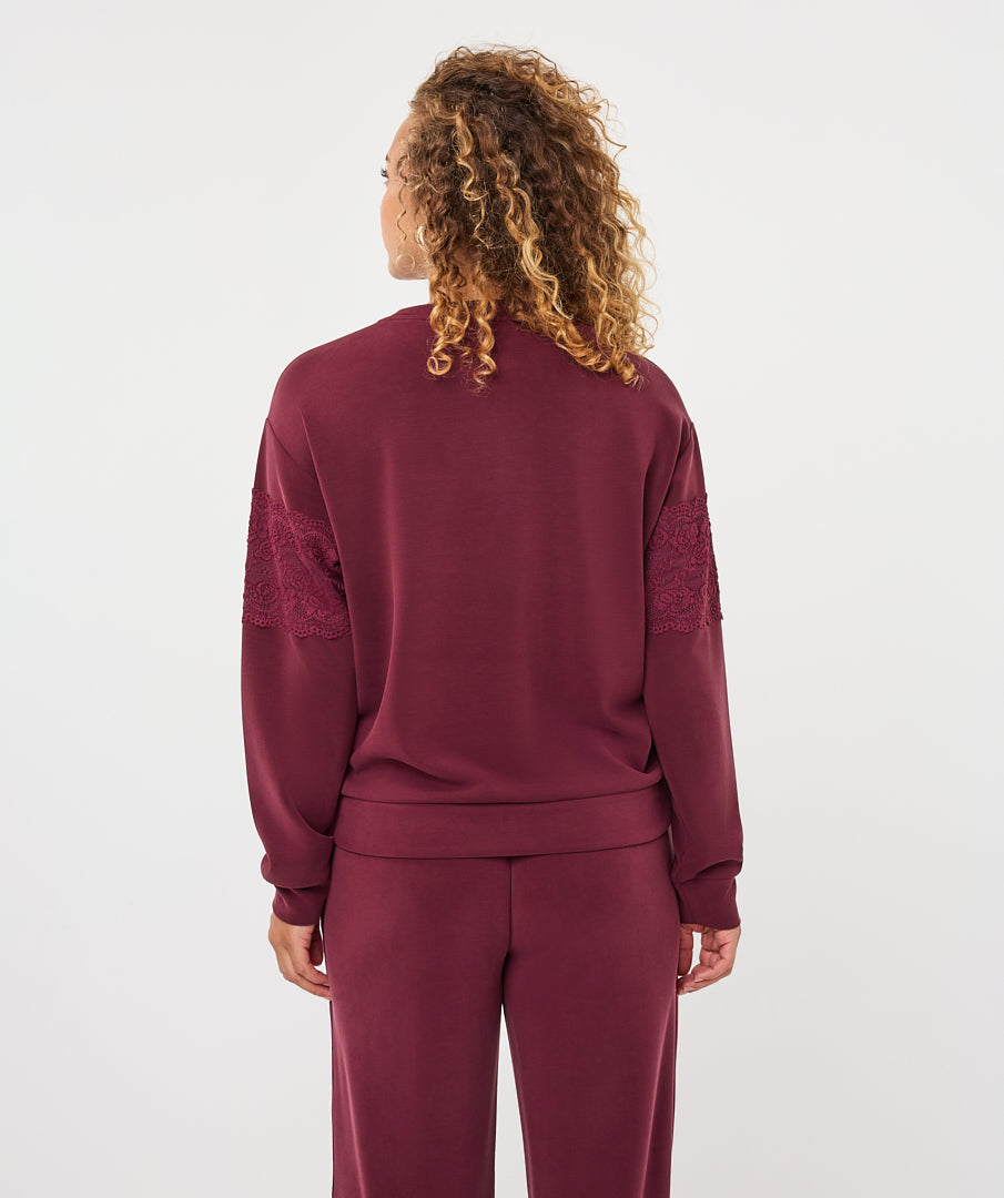 A woman viewed from the back wearing a matching burgundy lounge set, featuring a long-sleeve top with lace sleeve panels and matching pants, against a plain white studio background.