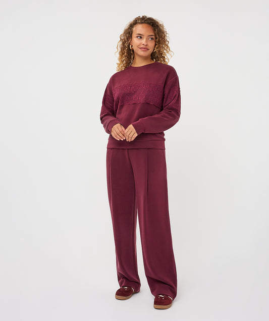 A woman modeling a matching burgundy lounge set consisting of a long-sleeve crewneck sweatshirt with tonal embroidery on the chest and matching wide-leg sweatpants, paired with burgundy slip-on shoes, against a plain light gray background.