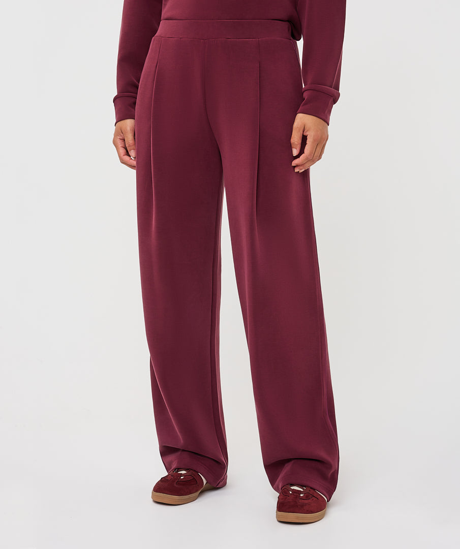 A person wearing high-waisted, wide-leg burgundy sweatpants with a matching long-sleeve top and burgundy slip-on shoes, shown from the waist down on a light gray studio background.