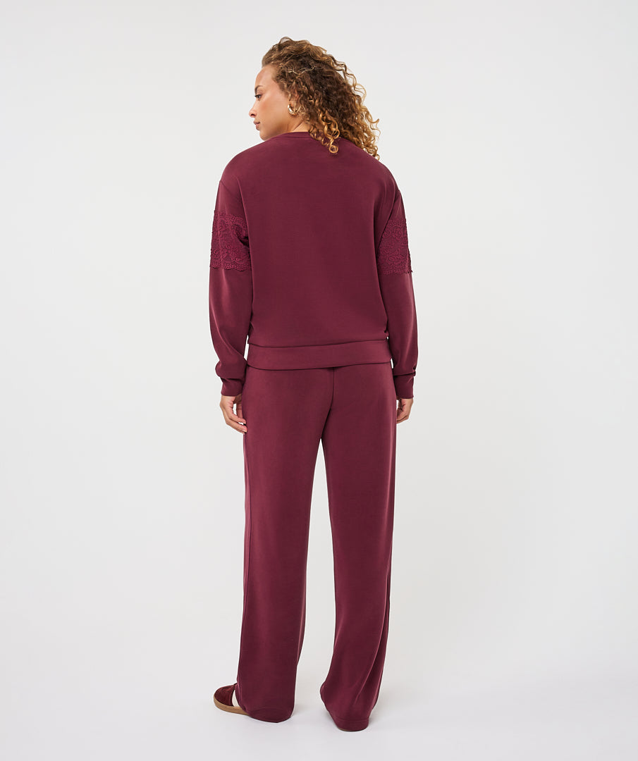 A woman with curly hair is shown from the back wearing a matching burgundy lounge set, consisting of a long-sleeve sweatshirt with textured shoulder panels and wide-leg sweatpants, against a plain white studio background.