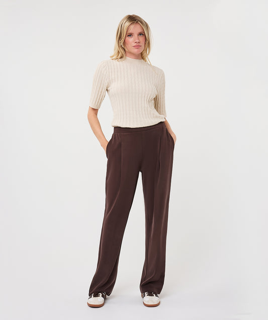 A woman modeling a cream ribbed short-sleeve mock-neck top and dark brown wide-leg trousers, standing against a light gray studio background.