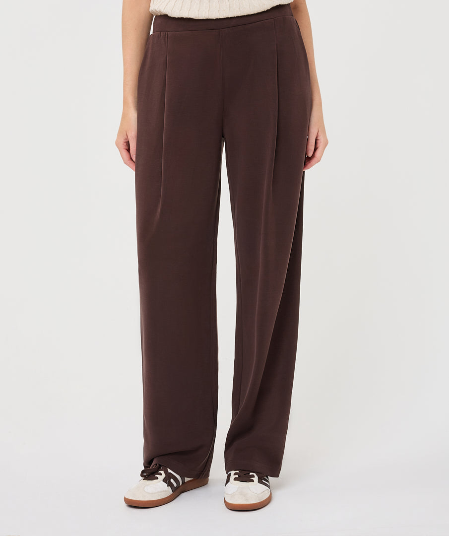 A person wearing dark brown wide-leg pleated trousers, styled with a cream knit top and white sneakers with black stripes, against a plain light gray studio background.
