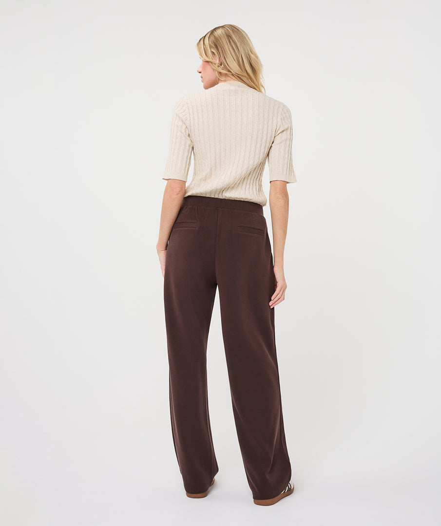 A woman is shown from behind wearing a cream ribbed short-sleeve mock-neck top and dark brown wide-leg trousers against a plain white studio background.