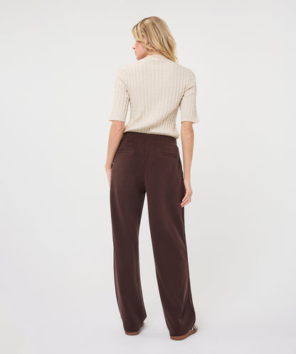 A woman is shown from behind wearing a cream ribbed short-sleeve mock-neck top and dark brown wide-leg trousers against a plain white studio background.