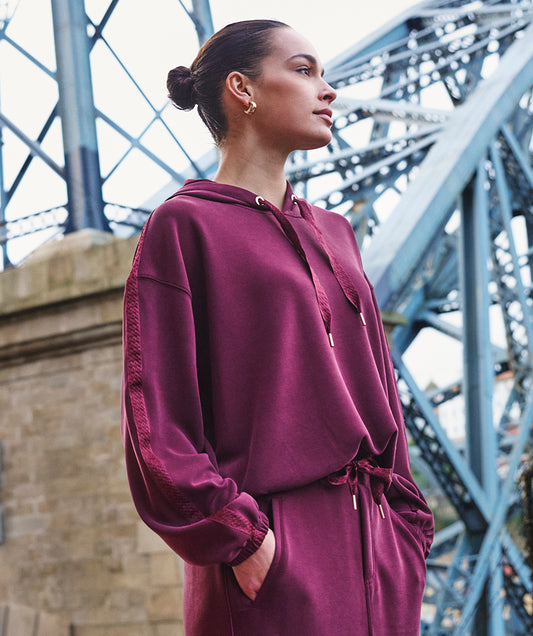 A woman wearing a matching burgundy lounge set with a hooded sweatshirt and drawstring skirt, standing in front of a blue steel bridge.