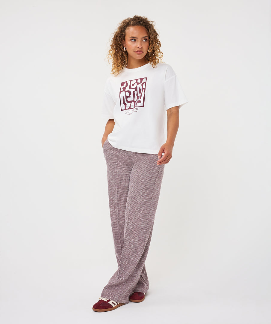 A woman modeling a white short-sleeve graphic t-shirt with a red and black block print, paired with dusty mauve wide-leg trousers and burgundy slide sandals, against a plain white studio background.