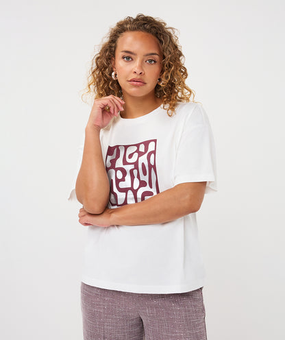 A woman with curly light brown hair wearing a white short-sleeve t-shirt with a maroon abstract graphic print on the front, paired with textured mauve pants, against a plain white studio background.