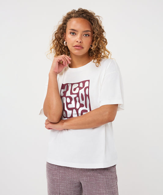 A woman with curly light brown hair wearing a white short-sleeve t-shirt with a maroon abstract graphic print on the front, paired with textured mauve pants, against a plain white studio background.