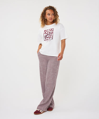 A woman modeling a white short-sleeve graphic t-shirt with a red and black block print, paired with dusty mauve wide-leg trousers and burgundy slide sandals, against a plain white studio background.
