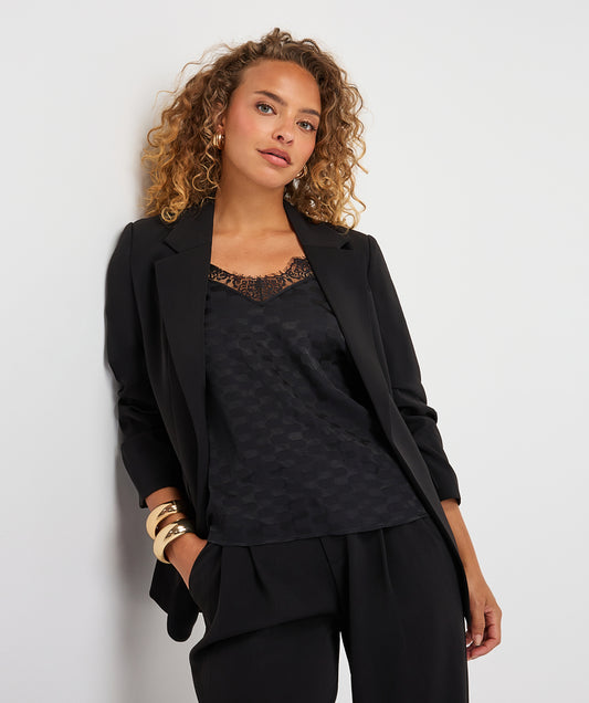 A woman with curly blonde hair models a black tailored blazer over a black lace-trim camisole and black trousers, accessorized with gold bracelets, against a plain light gray studio background.