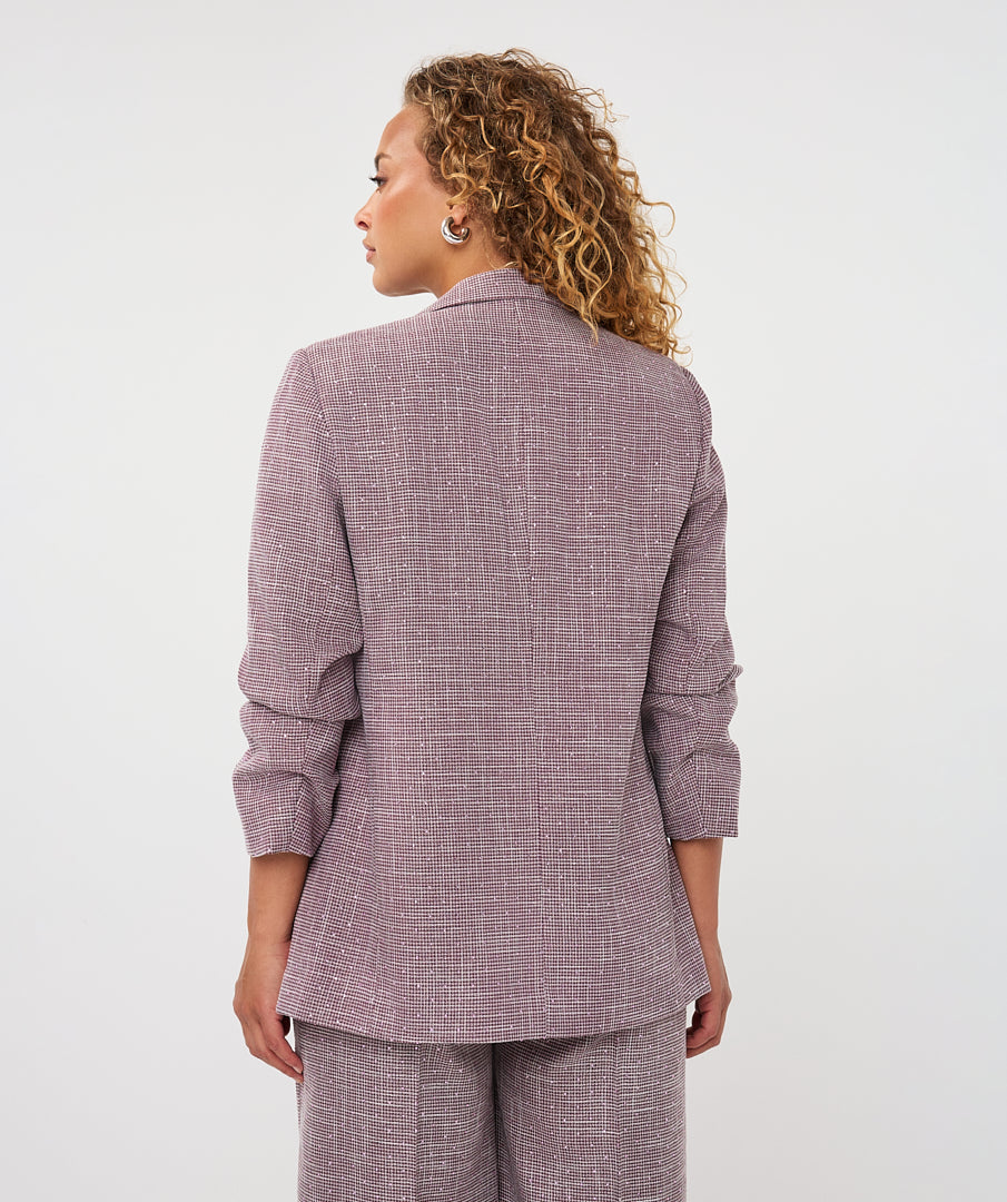 A woman with curly blonde hair is shown from the back wearing a textured mauve-purple blazer and matching trousers against a plain white studio background.