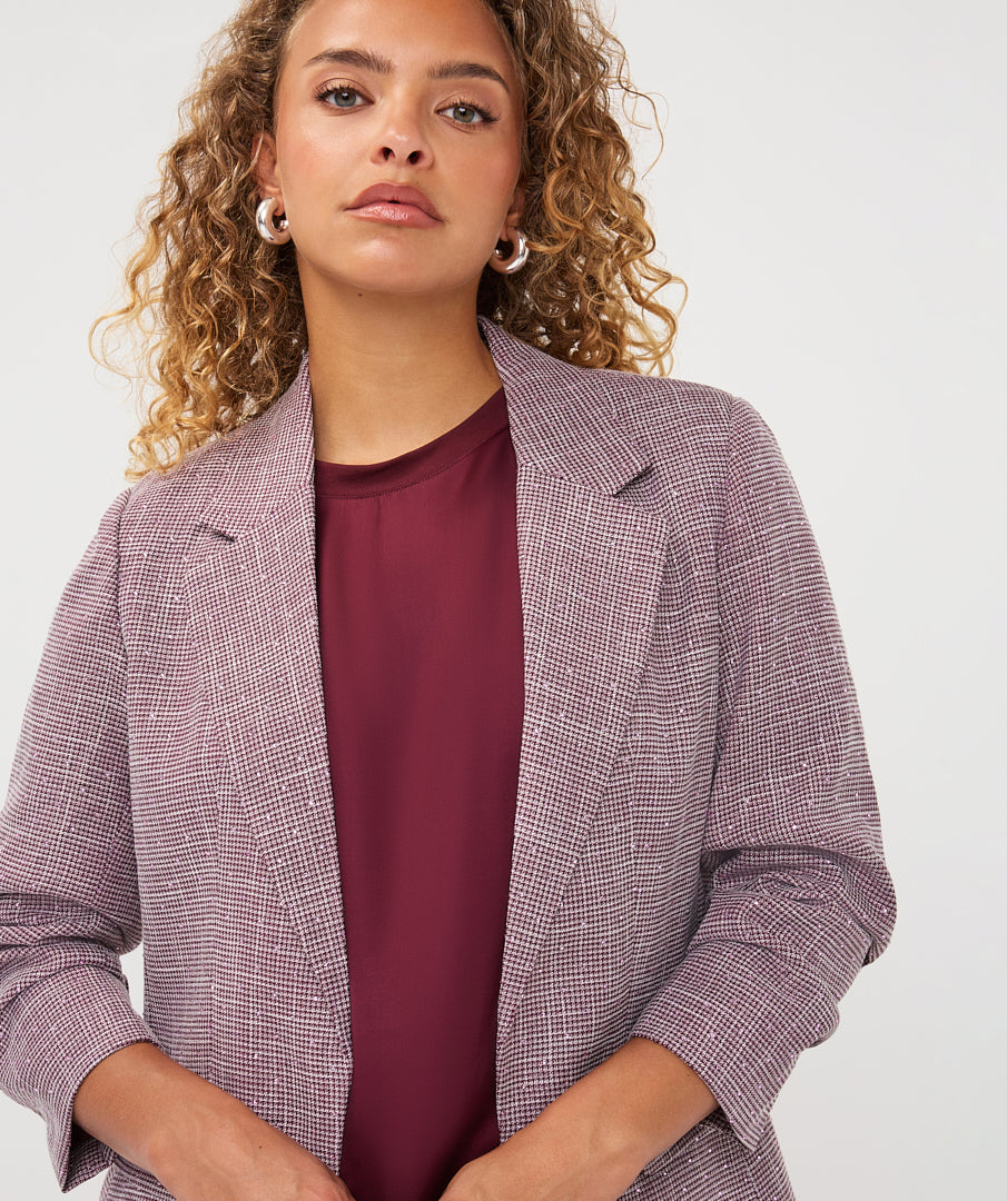 A woman with curly blonde hair wearing a textured, fine-checkered lavender and gray blazer over a deep burgundy top, photographed against a clean white studio background.